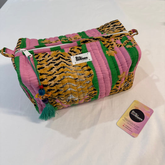 A quilted cosmetic pouch with a colorful striped pattern of pink, green, and tiger-print fabric, featuring a tassel zipper pull and a small white brand label. A pink and yellow tag is placed next to it on a white fabric surface.