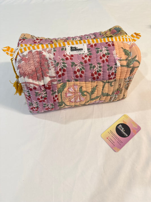 A quilted cosmetic pouch with a pink cherry and floral pattern, featuring yellow and white striped trim and tassels, displayed on a white fabric background next to a brand card.