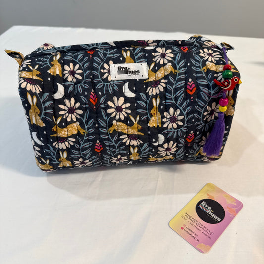 A quilted cosmetic pouch by Vera Bradley featuring a woodland print with rabbits, flowers, and leaves on a dark navy background. The pouch has a white brand label on the front and a colorful tassel charm on the side, displayed on a white surface next to a product tag.