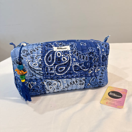 A blue quilted bandana-patterned cosmetic pouch with a tassel charm, shown on a white surface with its brand tag and a small card beside it.