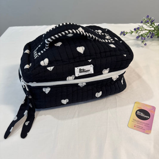 A black quilted cosmetic bag with a white heart pattern, featuring a top handle, zipper closure, and a small white brand label. The bag is displayed on a white fabric surface next to a small card and some purple flowers.