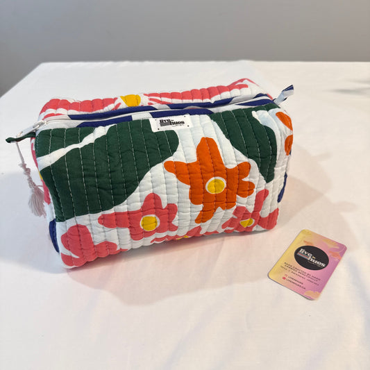 A quilted cosmetic pouch by Vera Bradley with a colorful floral pattern, placed on a white fabric surface next to its brand tag.