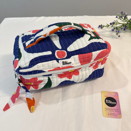 A quilted fabric cosmetic bag with a colorful abstract floral pattern in navy, white, coral, and green, shown on a white fabric surface next to a small card.