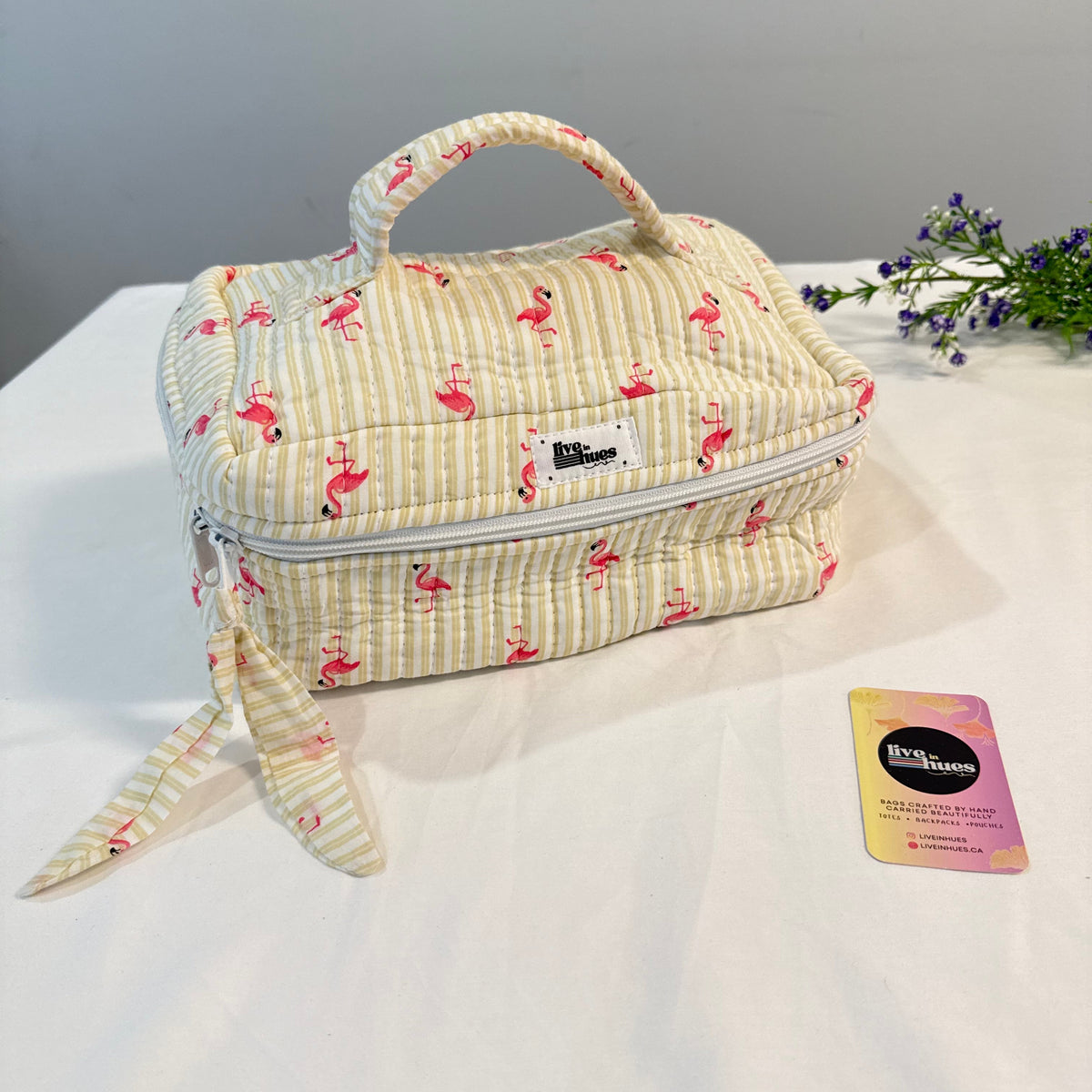 A quilted fabric toiletry bag with a top handle, featuring a cream and yellow striped pattern with pink flamingos. The bag has a white zipper and a matching fabric wristlet strap.