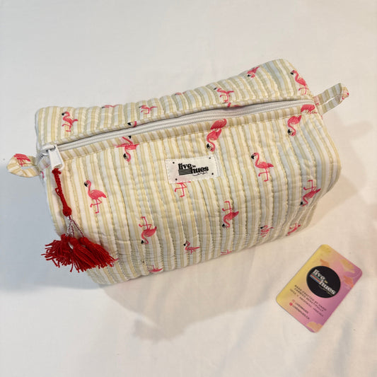 A quilted cosmetic pouch with a cream and light blue striped pattern, decorated with pink flamingos, featuring a red tassel zipper pull and a white brand label. A small pink and yellow tag is placed next to it on a white fabric background.