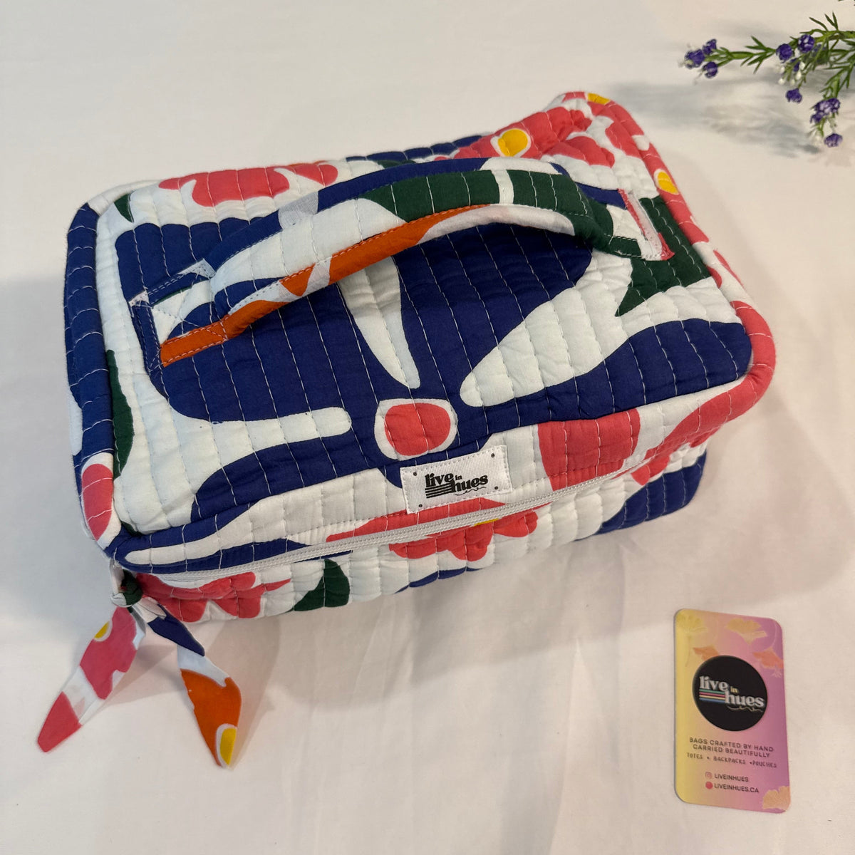 A quilted fabric cosmetic pouch with a colorful abstract print in navy, white, pink, orange, and green, displayed on a white fabric surface next to a small branded card.