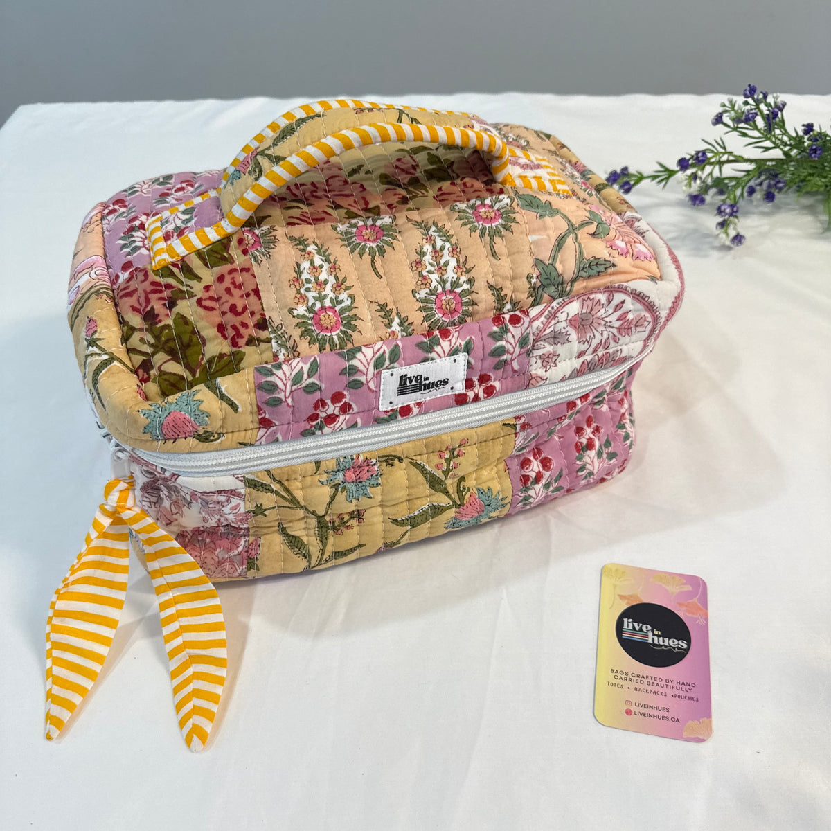 A quilted patchwork cosmetic bag with a floral pattern in pink, yellow, and green, featuring a striped yellow and white handle and matching ribbon ties. A small card for 'The Beauty Box' is placed next to it on a white fabric surface.