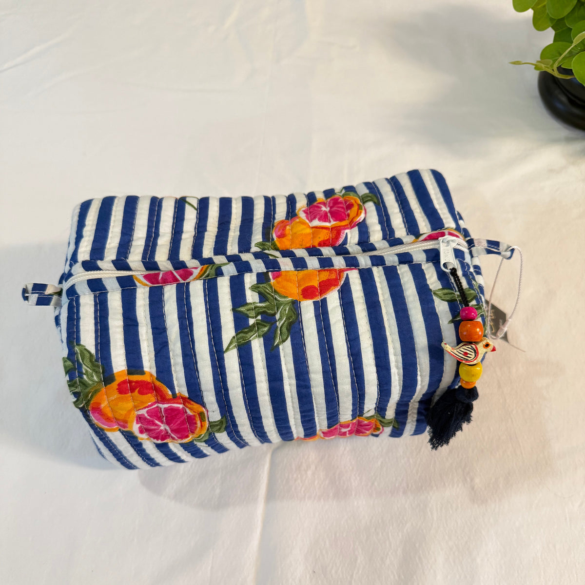 A fabric pouch with a navy blue and white striped pattern, decorated with colorful citrus fruit prints in orange, pink, and yellow. The pouch features a zippered top closure and a decorative tassel charm with wooden beads on the side, resting on a white fabric background.