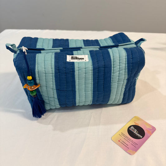 A quilted cosmetic pouch with vertical stripes in navy, medium blue, and light blue, featuring a tassel charm and a zip closure. A small brand tag and a colorful card are placed next to it on a white fabric surface.
