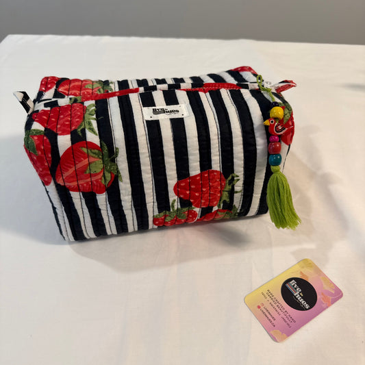 A fabric cosmetic pouch with a black and white striped pattern and red strawberry motifs, featuring a green tassel and a small card next to it on a white surface.