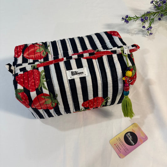 A fabric cosmetic pouch with a black and white striped pattern on one side and red strawberries on the other, featuring a green tassel charm and a small white brand label. A small card is placed next to it on a white fabric background.