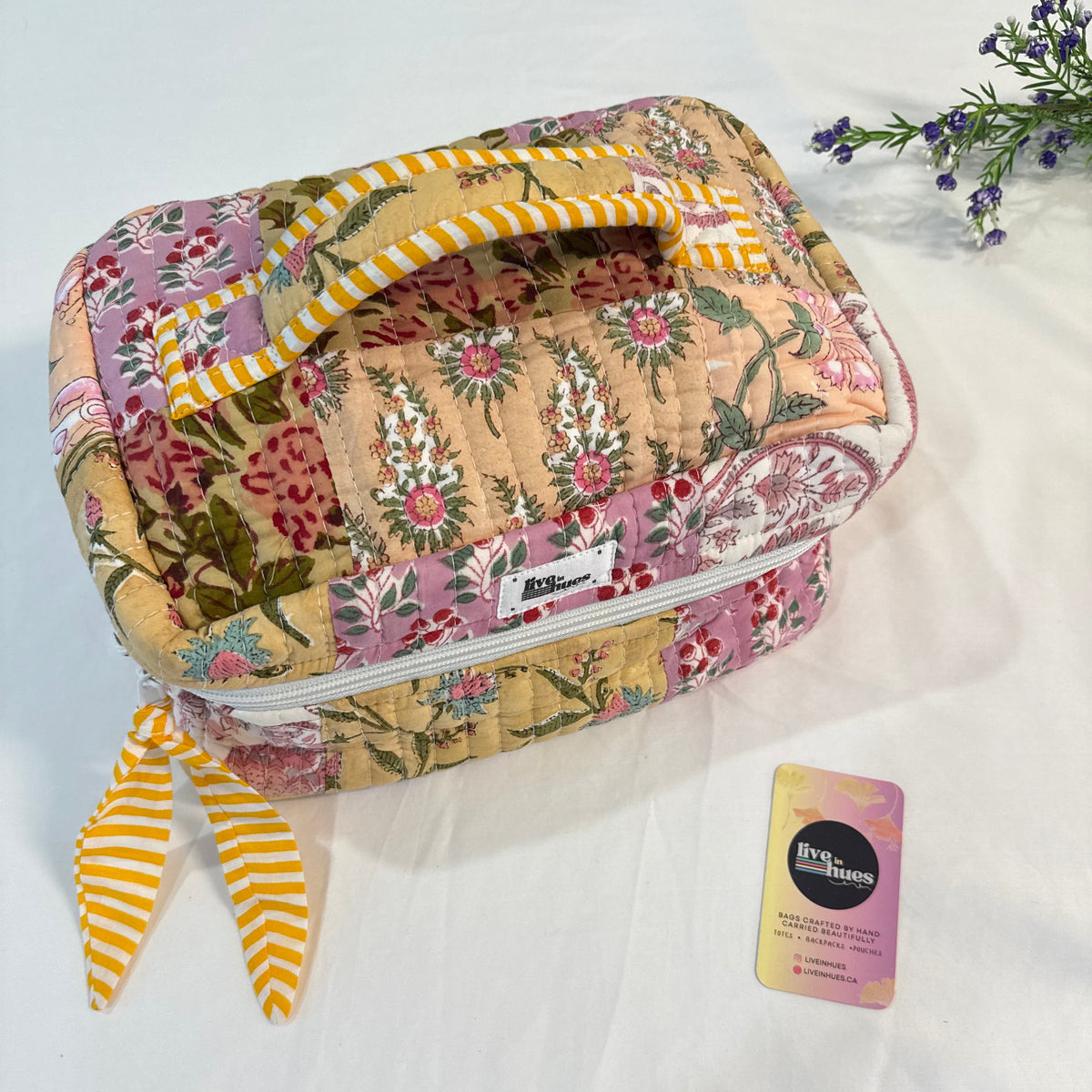 A quilted patchwork cosmetic bag with a floral pattern and yellow-and-white striped handles, shown on a white fabric background with a small card and decorative flowers.