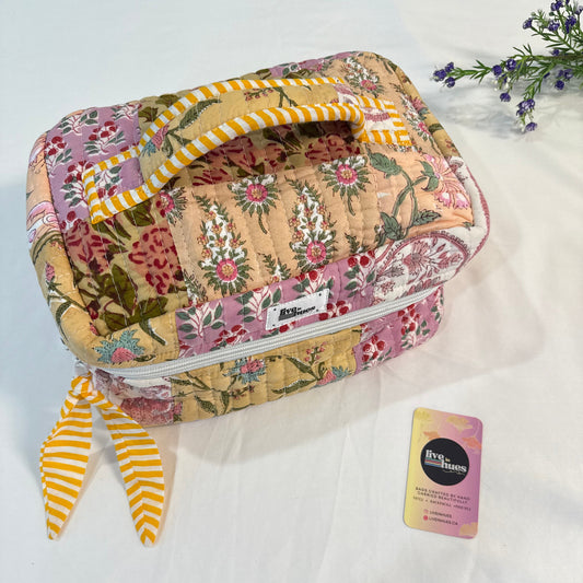A quilted patchwork cosmetic bag with a floral pattern and yellow-and-white striped handles, shown on a white fabric background with a small card and decorative flowers.