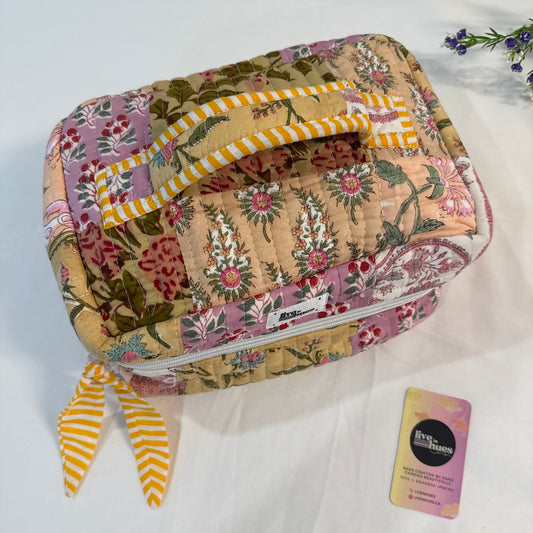 A quilted fabric cosmetic bag with a multicolor floral and botanical print, featuring a yellow and white striped handle and matching ribbon tie. A small brand card is placed next to it on a white fabric background.