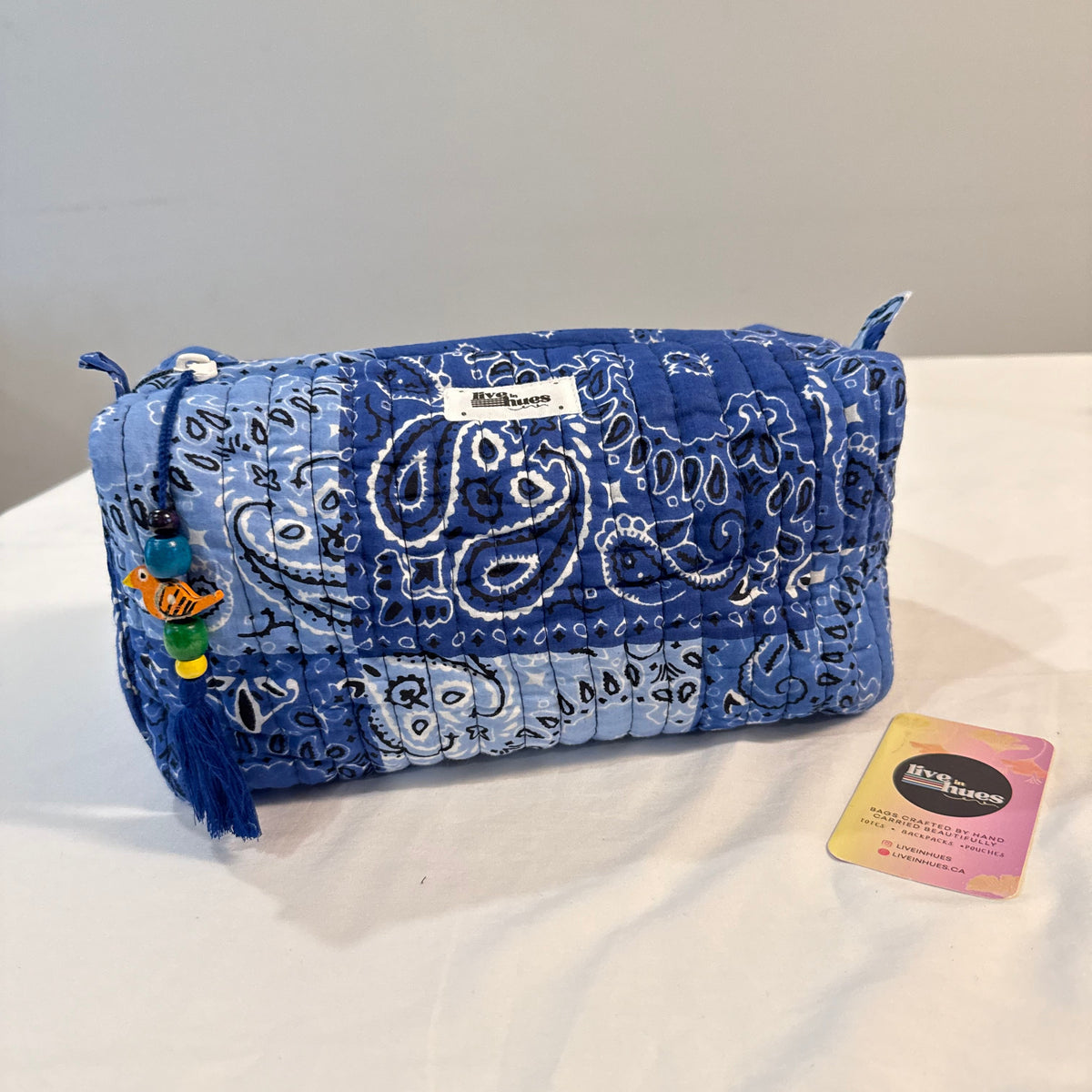 A blue quilted bandana-patterned cosmetic pouch with a tassel charm, shown on a white surface with its brand tag and a small card beside it.