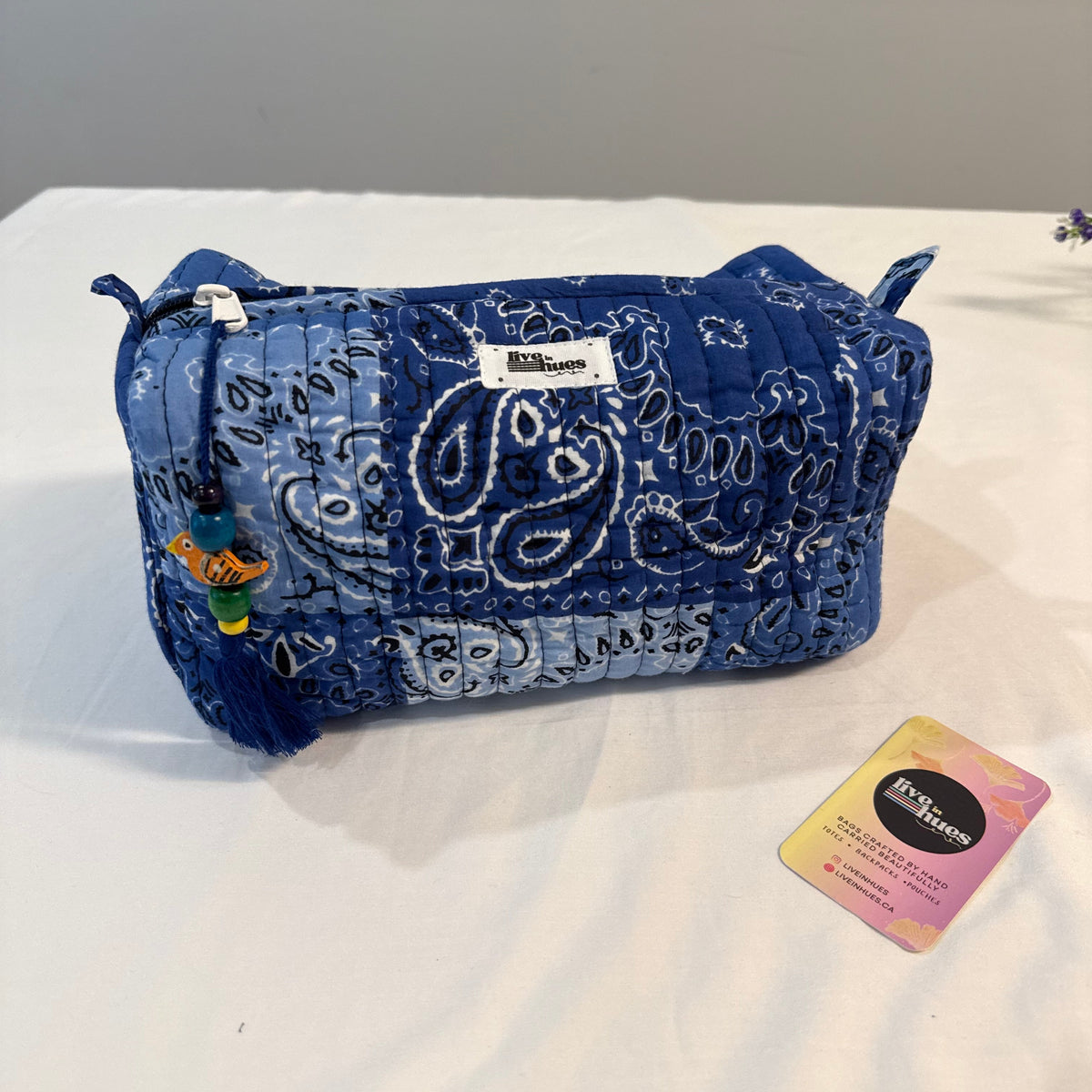 A blue quilted cosmetic pouch with a white paisley bandana print, featuring a small tassel charm and a white brand label that reads 'Briarwood'. A pink and yellow Briarwood gift tag lies next to it on a white fabric surface against a gray background.
