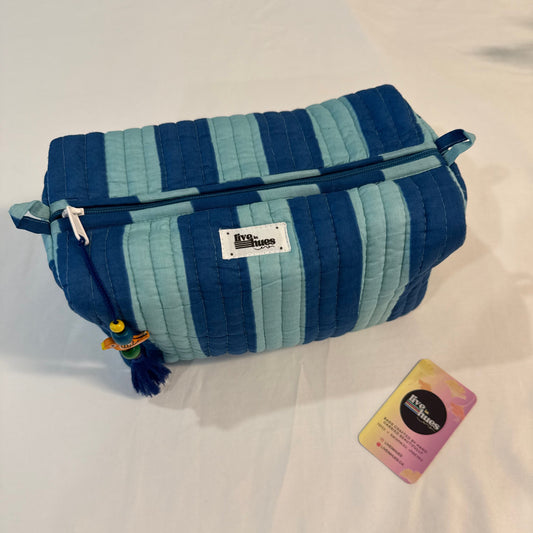 A quilted cosmetic pouch with vertical stripes in navy blue, medium blue, and light blue, featuring a white brand label and a decorative tassel zipper pull. It is displayed on a white fabric background next to a small card.