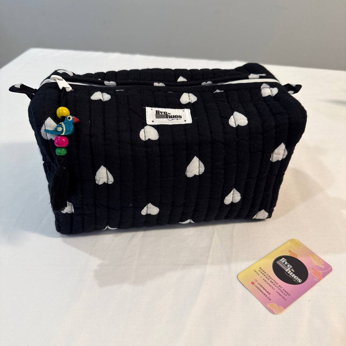 A black quilted cosmetic pouch with a white heart pattern, featuring a colorful bird charm and a white brand label, displayed on a white surface next to a small card.