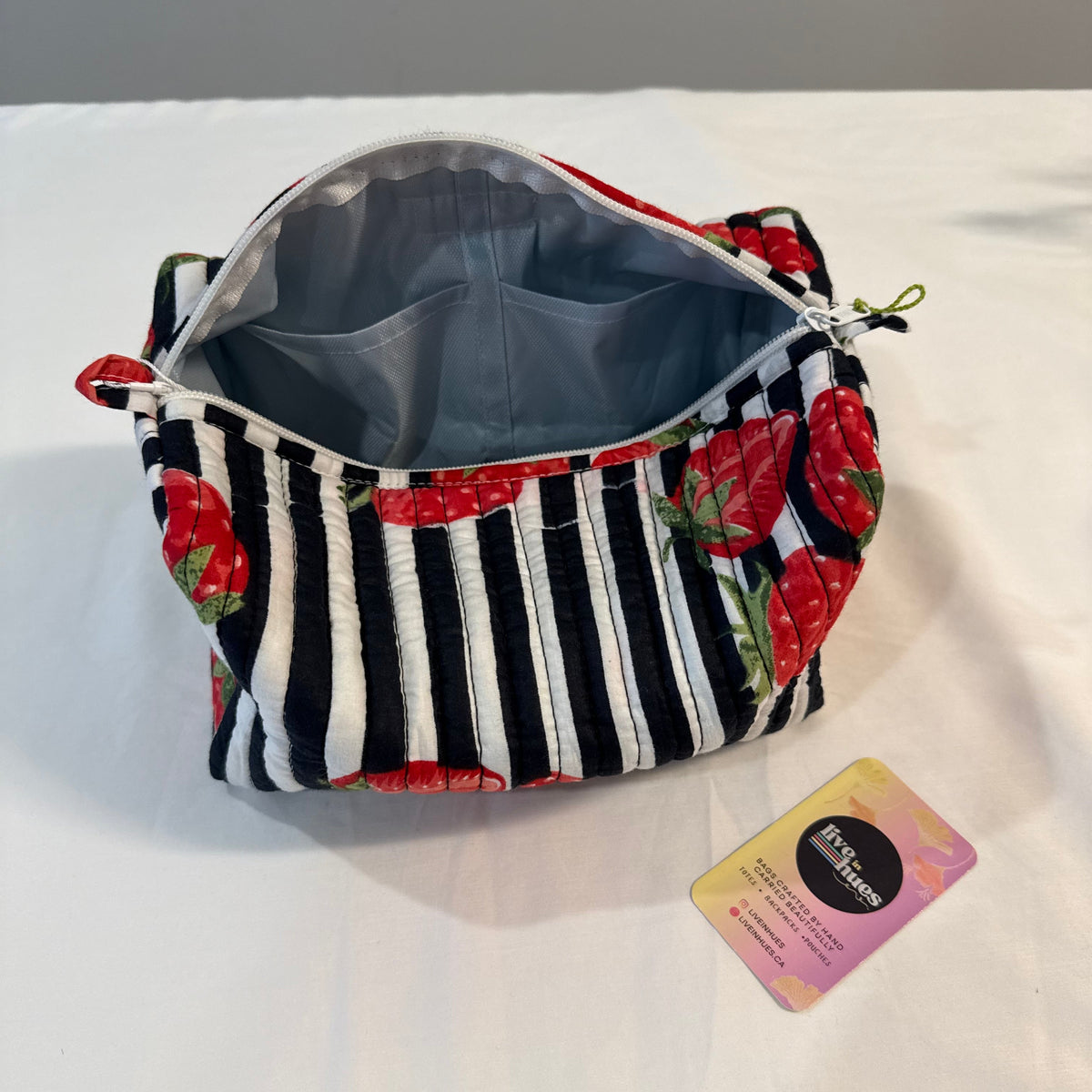A zippered cosmetic pouch with a black and white striped pattern and red strawberry motifs, shown open on a white fabric surface with a small card beside it.