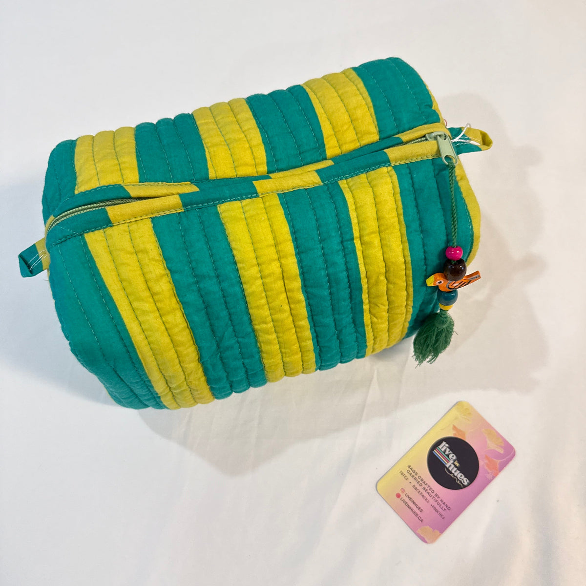 A quilted cosmetic pouch with alternating teal green and yellow stripes, featuring a gold zipper and a small hula dancer charm. A small brand card is placed next to it on a white fabric background.