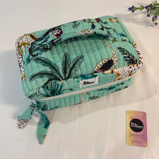 A quilted cosmetic bag with a tropical print in mint green, featuring palm leaves, flamingos, and birds, shown on a white fabric background next to a small card.