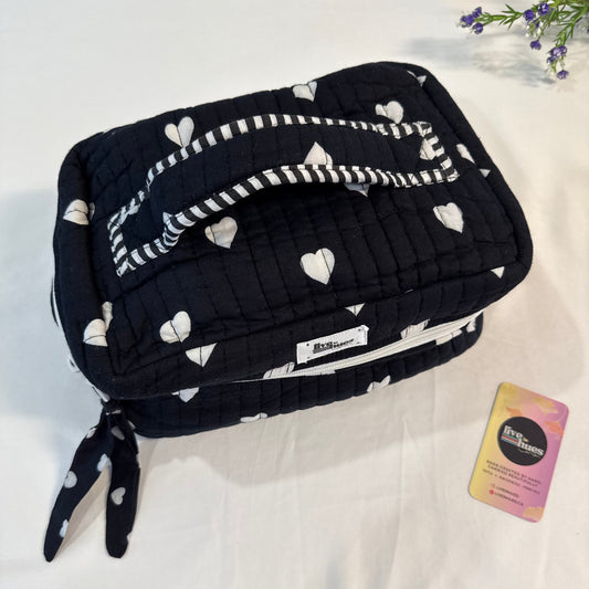 A quilted black cosmetic bag with a white heart and stripe pattern, featuring a top handle and a matching ribbon tie, displayed on a white fabric background next to a small card.