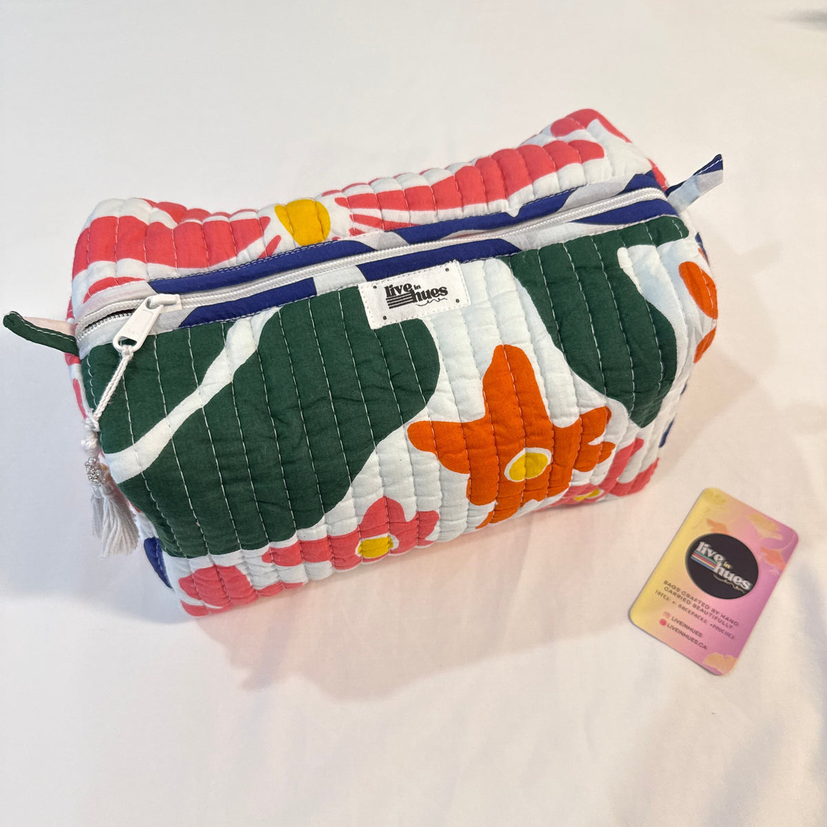 A quilted cosmetic pouch with a colorful floral pattern in green, orange, pink, and white, featuring a blue zipper. A small white label and a pink gift card are placed next to it on a white fabric background.