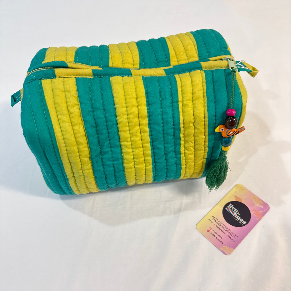 A quilted cosmetic pouch with vertical teal and yellow stripes, featuring a green tassel charm with a small figurine and a pink and yellow tag, displayed on a white fabric background.