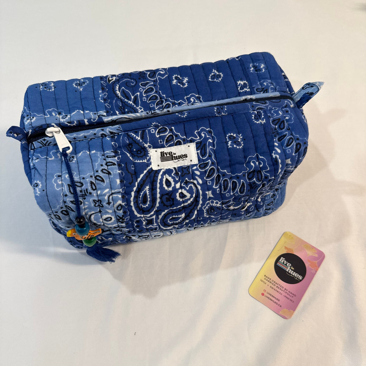 A blue and white paisley bandana-patterned quilted cosmetic pouch with a zipper closure, shown on a white fabric background next to a small pink and yellow tag.