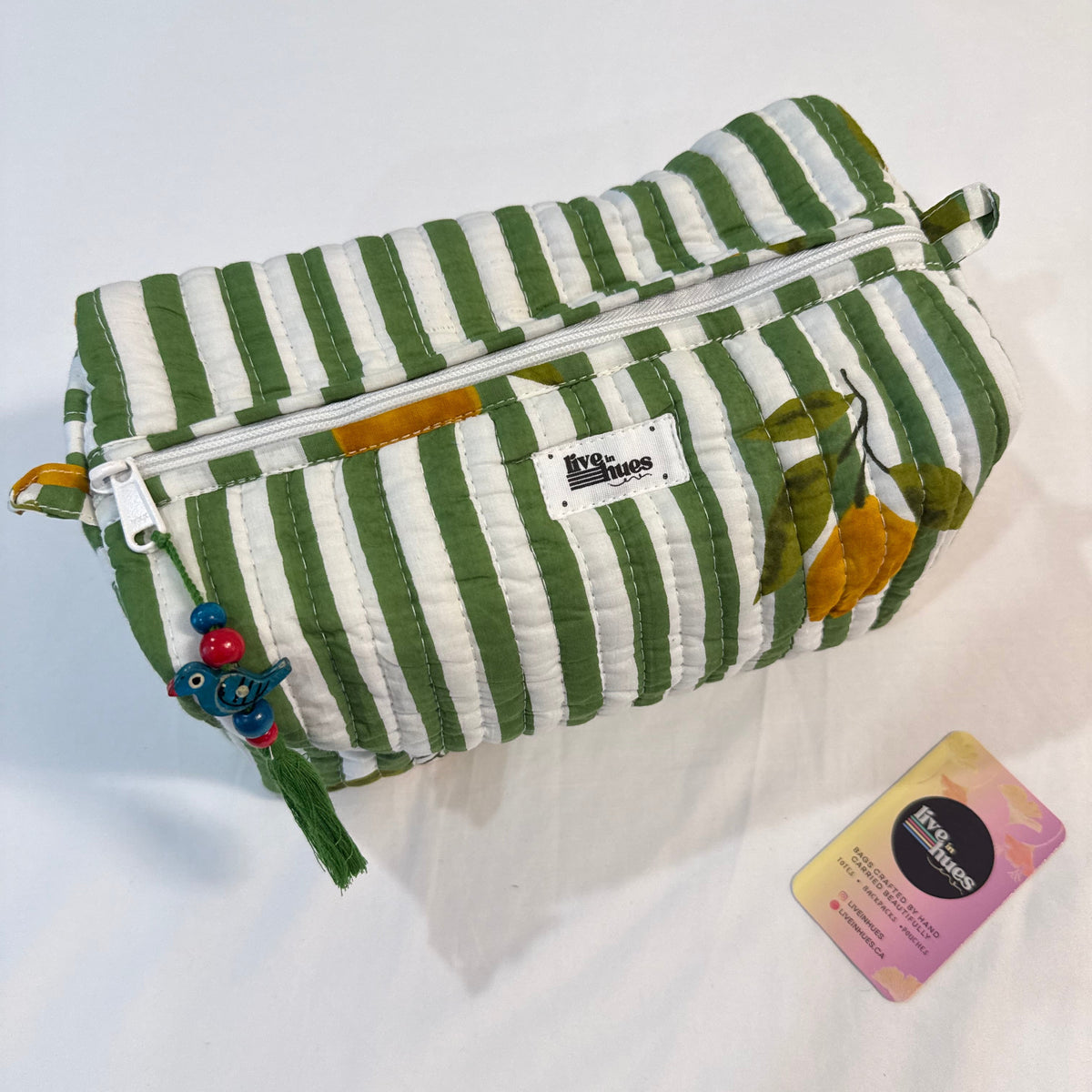 A green and white striped fabric pouch with a zipper closure, featuring a small white brand label and colorful hanging tassels, displayed on a white fabric background next to a small card.
