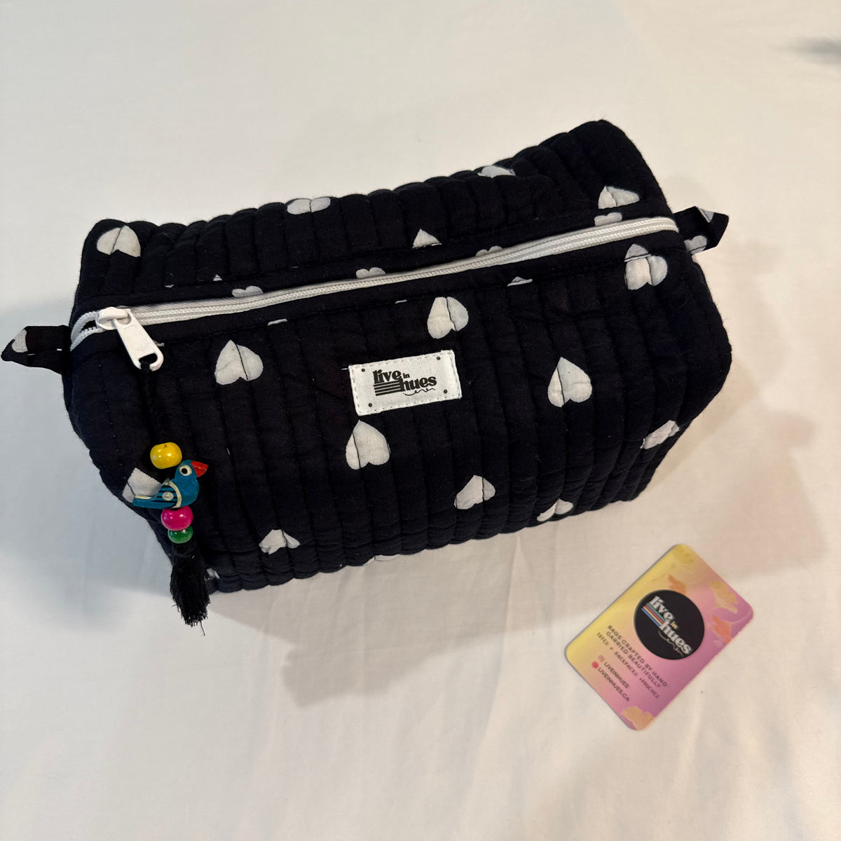 A black quilted cosmetic pouch with a white heart pattern, featuring a silver zipper, a white brand label, and a small colorful charm, displayed on a white fabric surface next to a small card.