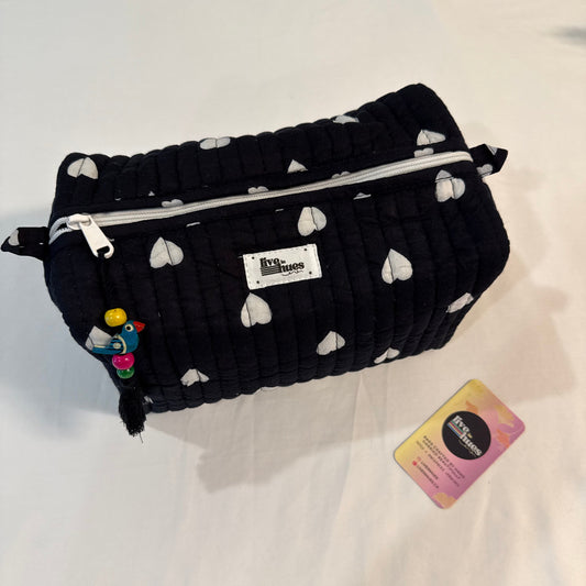 A black quilted cosmetic pouch with a white heart pattern, featuring a silver zipper, a white brand label, and a small colorful charm, displayed on a white fabric surface next to a small card.