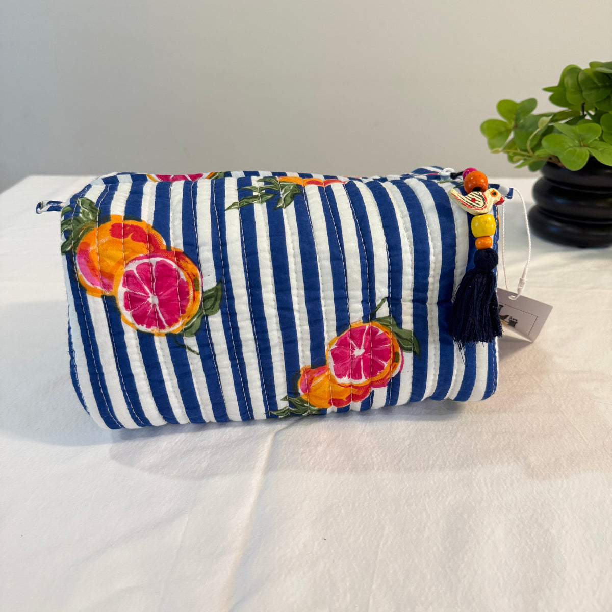 A fabric cosmetic pouch with navy and white vertical stripes and colorful citrus fruit prints, featuring a black tassel charm with a cartoon character, resting on a white fabric surface.
