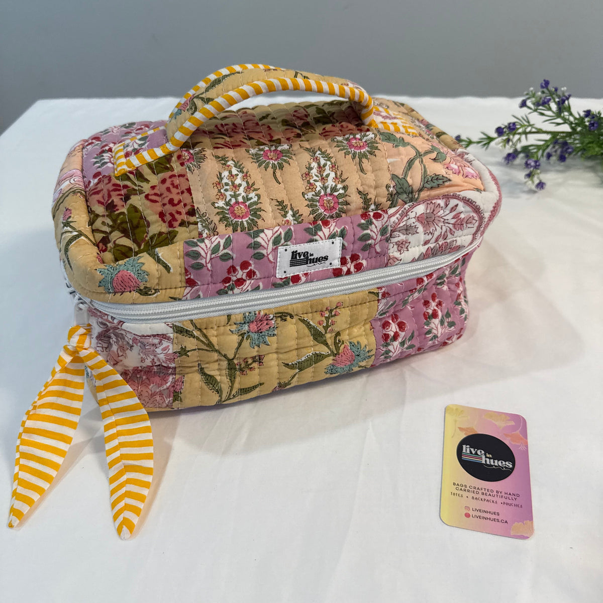 A quilted patchwork cosmetic bag with a floral pattern in pink, yellow, and green, featuring a striped yellow-and-white handle and matching tie strap. A small white label on the front reads 'Blossom'.
