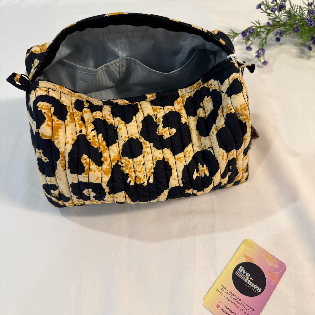 A quilted cosmetic bag with a black and mustard yellow leopard print pattern, shown open on a white fabric surface next to a brand card.