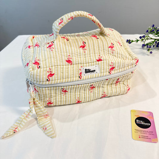 A quilted travel toiletry bag with a top handle, featuring a cream and yellow stripe pattern with pink flamingo prints. The bag has a white zipper, a matching fabric wristlet strap, and a small white brand label that reads 'Himiko'.