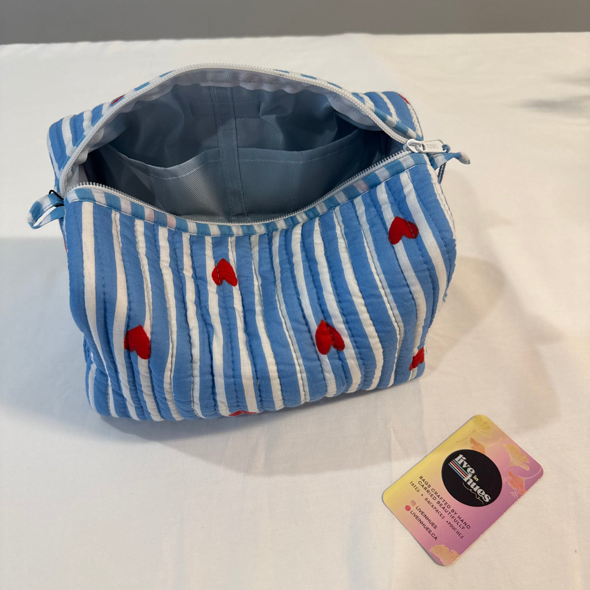 A blue and white striped fabric pouch with red heart patterns, shown open on a white surface with a small card next to it.